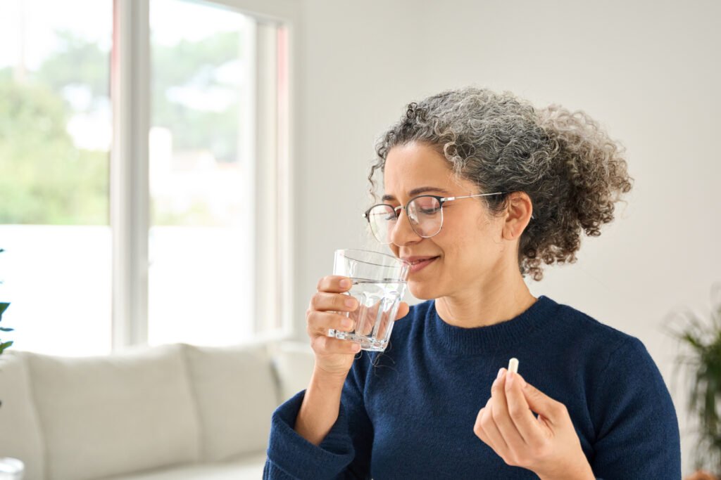 Suplementación en la menopausia: Mujer sana y feliz de mediana edad, sentada en casa, tomando una pastilla y sosteniendo un vaso de agua. Mujer madura de 50 años tomando suplementos vitamínicos para su salud, concepto de tratamiento nutricional diario por la mañana.