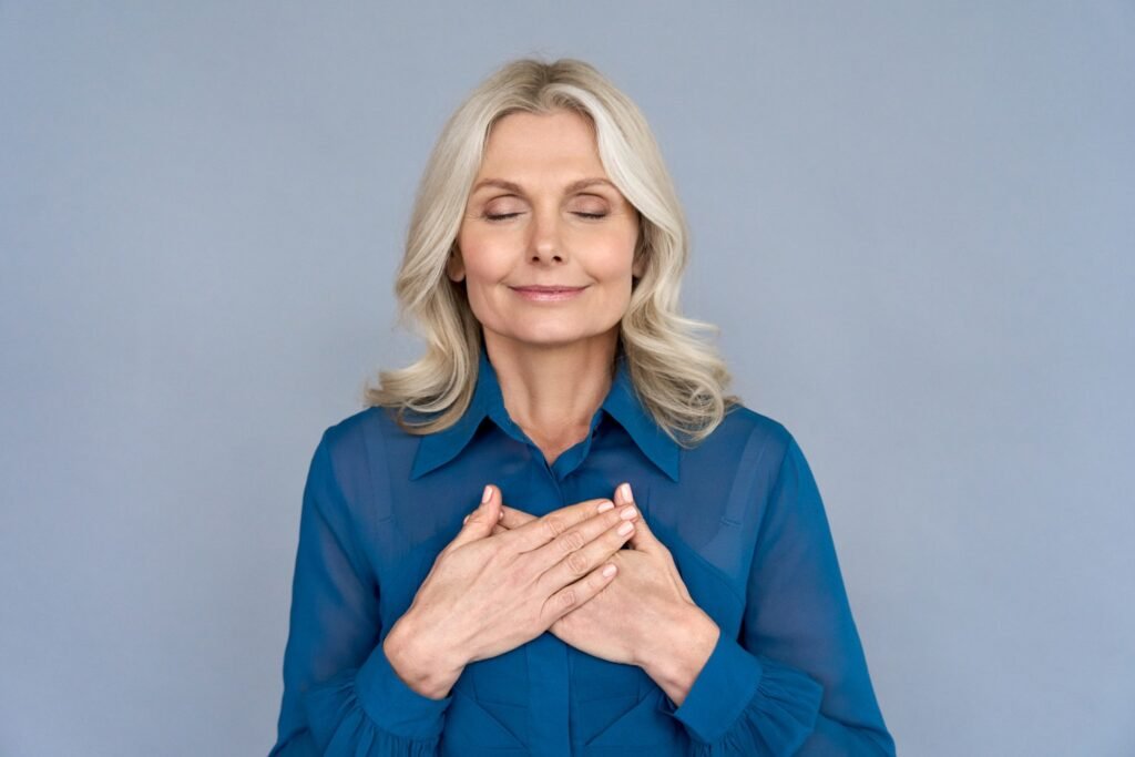 Conexión Interior en Menopausia. Mujer de mediana edad feliz, consciente y agradecida, con las manos sobre el pecho, meditando con los ojos cerrados, aislada sobre fondo gris, sintiendo ausencia de estrés, gratitud, equilibrio de salud mental, concepto de paz mental.